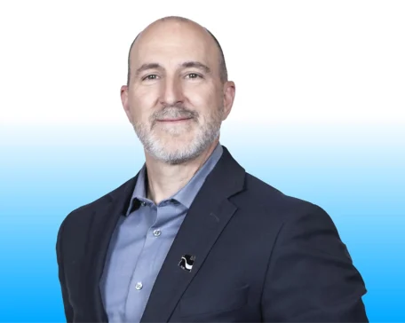 Bald man with a beard wearing a suit jacket over a blue shirt, smiling against a white and blue gradient background.