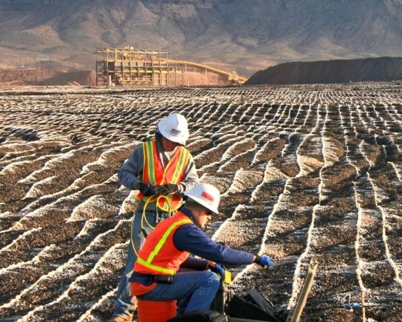 Engineers in safety gear conduct tests on a rocky field with grid lines, mountains in the background.