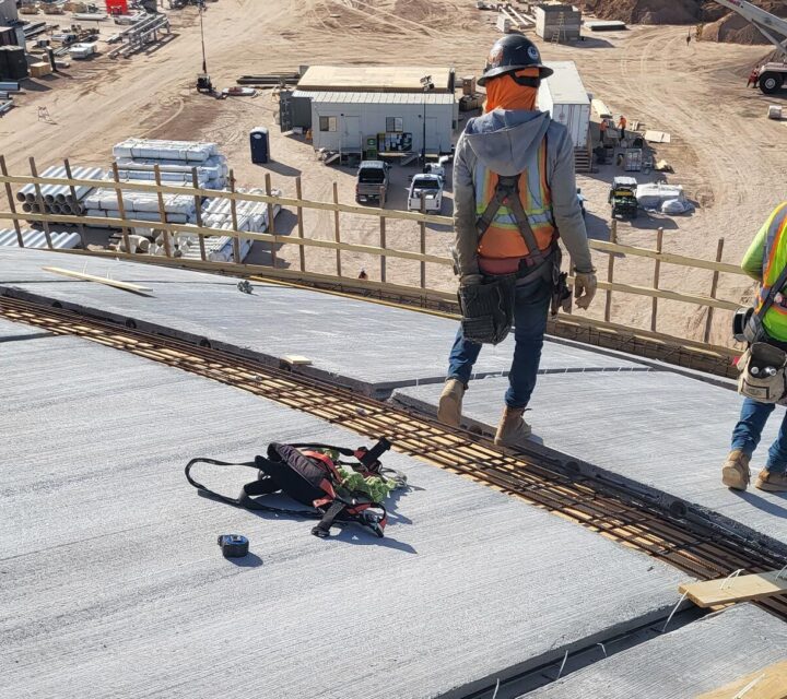 Construction workers in safety gear stand on a curved concrete surface at a work site.
