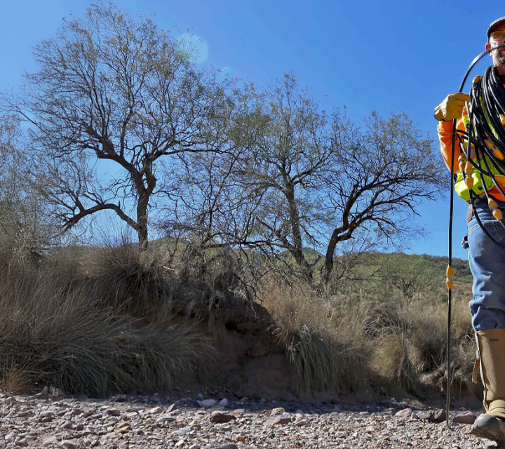 Worker in high-visibility gear carries coiled cables in a rocky, dry landscape with sparse trees.