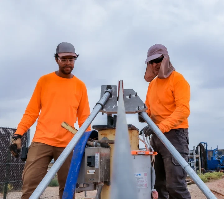 Two workers in orange shirts operate machinery to cut metal pipes outdoors.