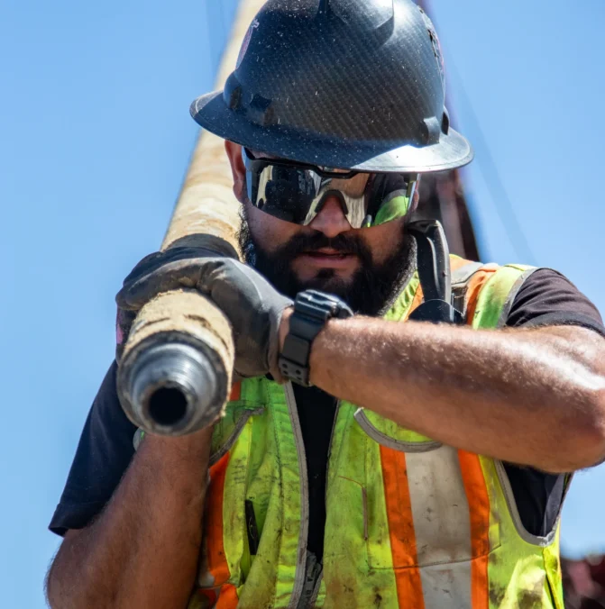 Construction worker in safety gear and sunglasses, holding a large pipe at a work site.