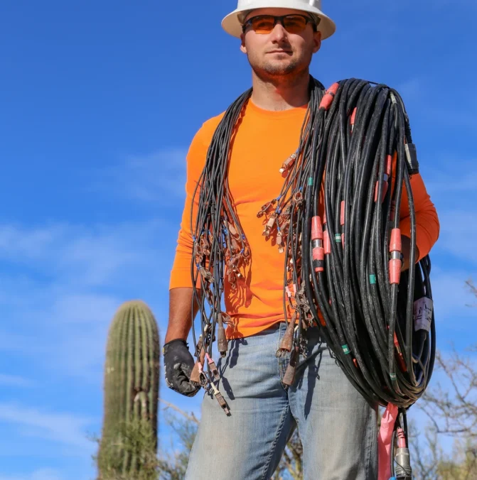 Worker in hard hat carrying cables with a cactus and blue sky in the background.