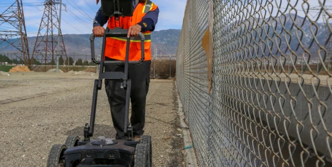Worker in safety gear uses ground-penetrating radar near a chain-link fence on a clear day.