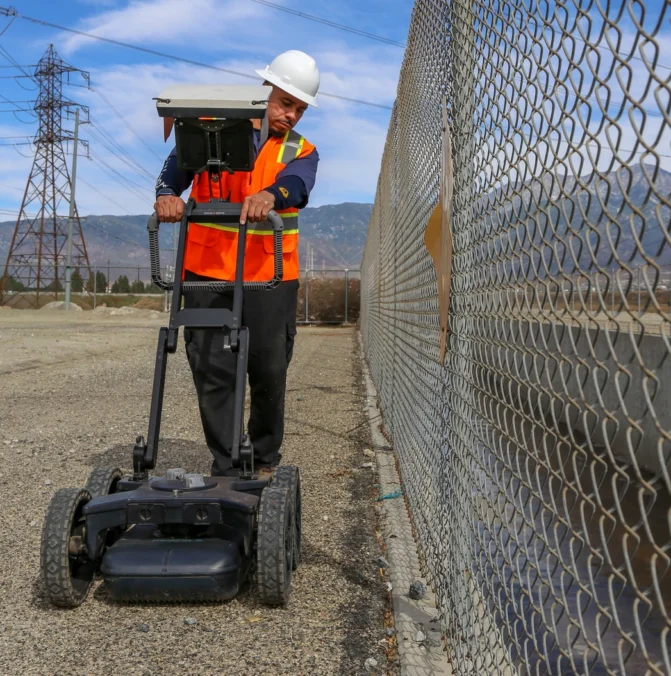 Worker in safety gear uses ground-penetrating radar near a chain-link fence on a clear day.
