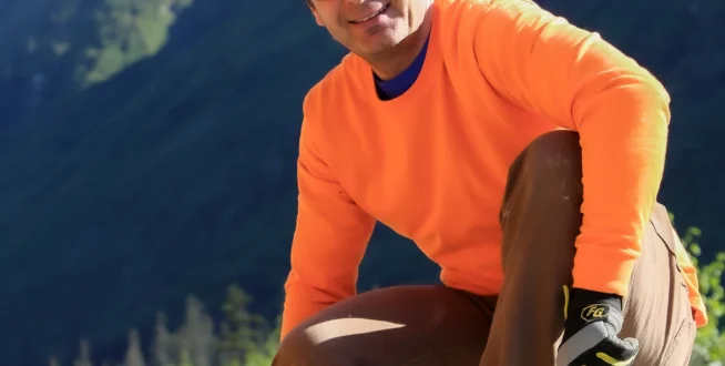 Worker in orange long-sleeve and hard hat kneels on gravel, with snowy mountains in background.