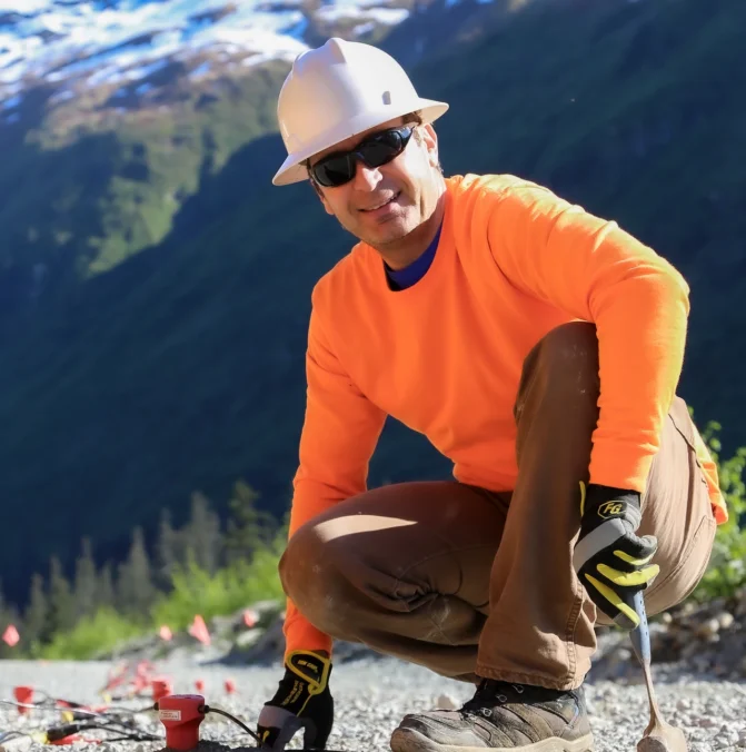 Worker in orange long-sleeve and hard hat kneels on gravel, with snowy mountains in background.