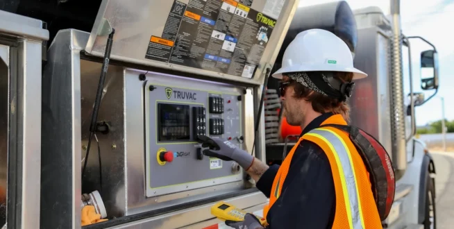 Worker in safety gear operates controls on industrial machinery with open panel outdoors.
