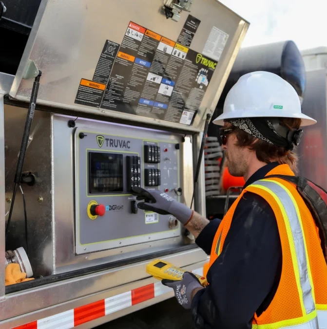 Worker in safety gear operates controls on industrial machinery with open panel outdoors.