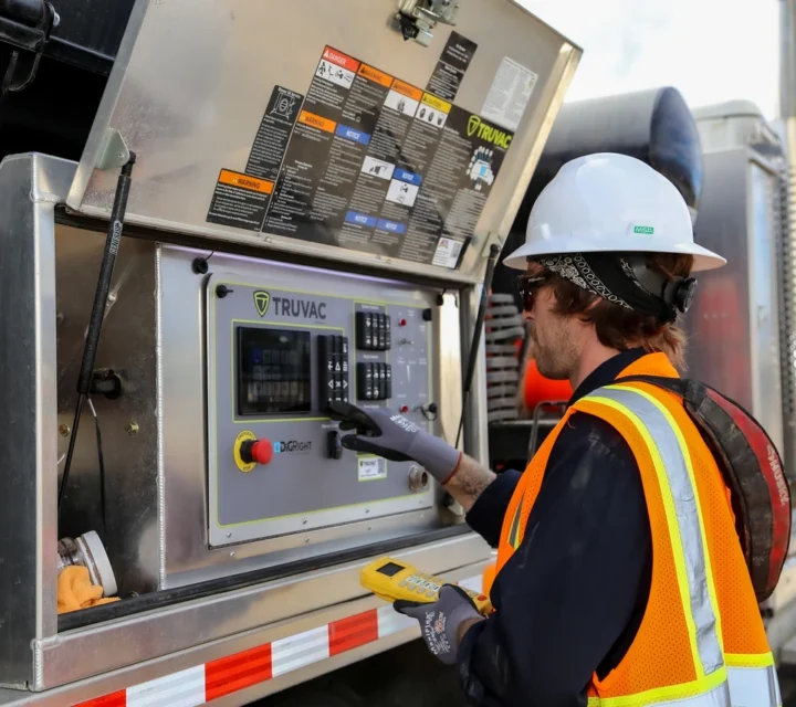 Worker in safety gear operates controls on industrial machinery with open panel outdoors.