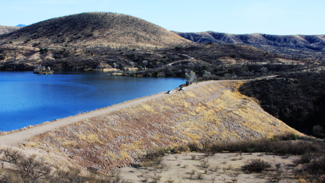 A serene hilly landscape with a blue lake, dirt path, and parked cars under a clear sky.