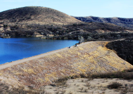 A serene hilly landscape with a blue lake, dirt path, and parked cars under a clear sky.