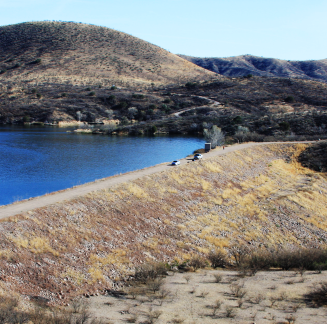 A serene hilly landscape with a blue lake, dirt path, and parked cars under a clear sky.