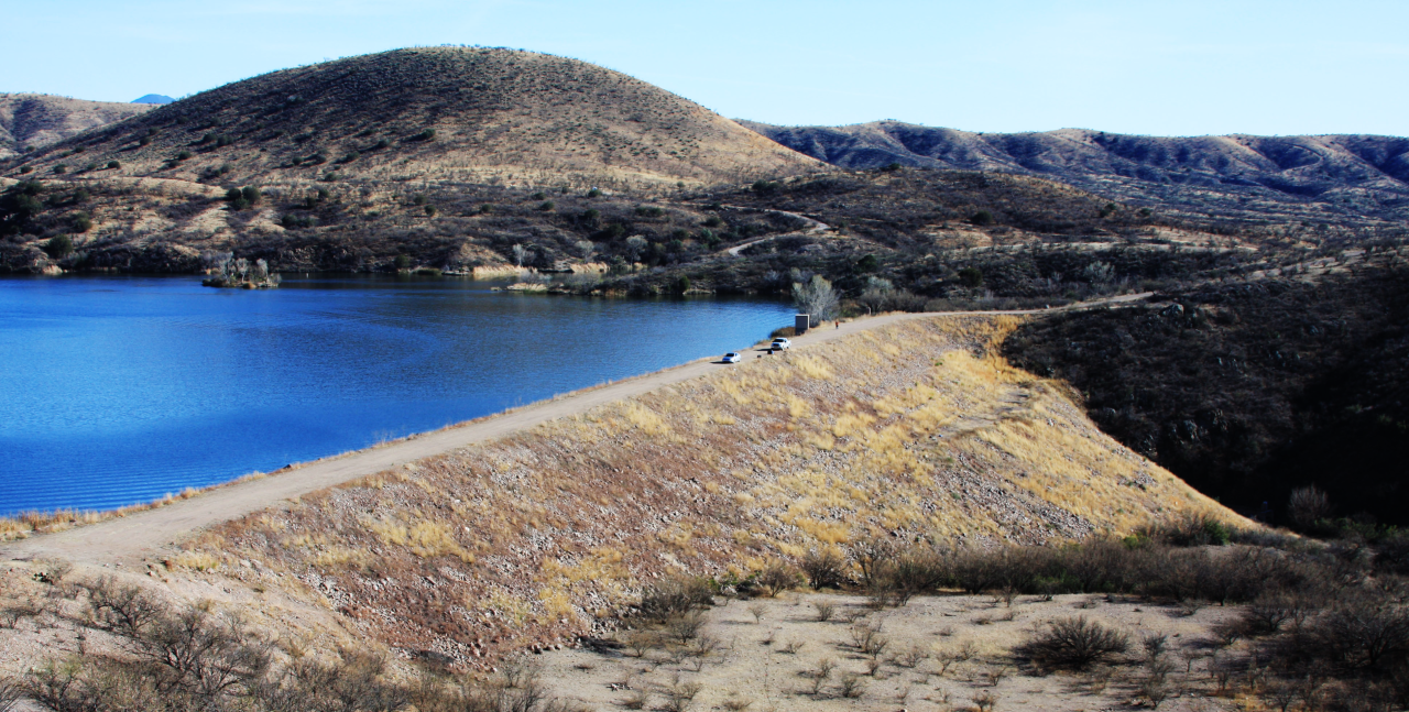 A serene hilly landscape with a blue lake, dirt path, and parked cars under a clear sky.