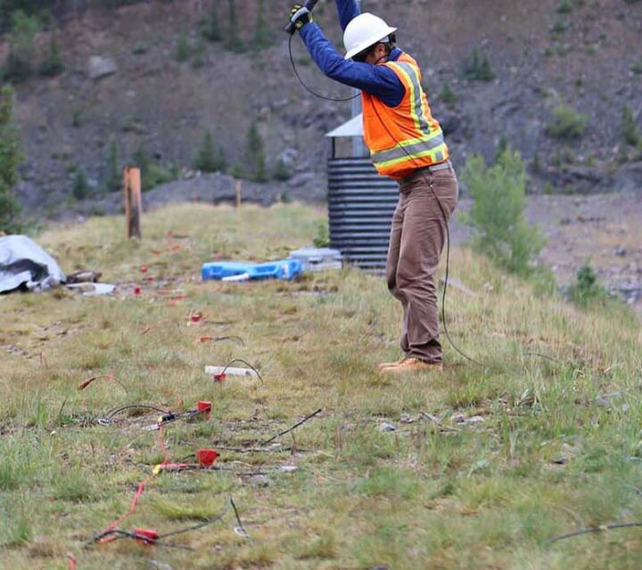 Person in safety gear conducting geophysical survey on grassy hillside.