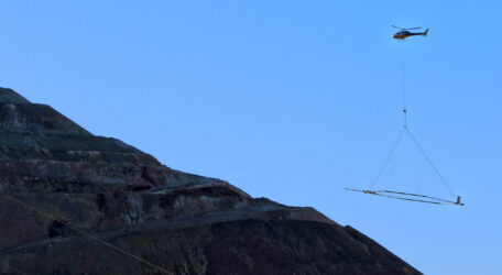 Helicopter lifting equipment over a rugged, stepped landform against a clear blue sky.