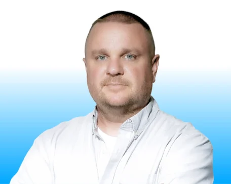 Man with short hair and a beard wearing a white shirt, standing against a blue gradient background.