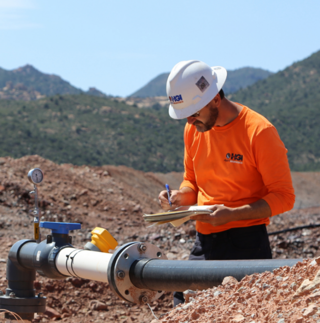 Engineer inspecting pipeline in a rugged mountain landscape, taking notes on a clipboard.