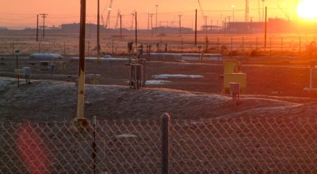 Sunset over industrial site with cranes, fenced area, and utility boxes in the foreground.