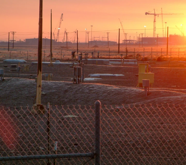 Sunset over industrial site with cranes, fenced area, and utility boxes in the foreground.