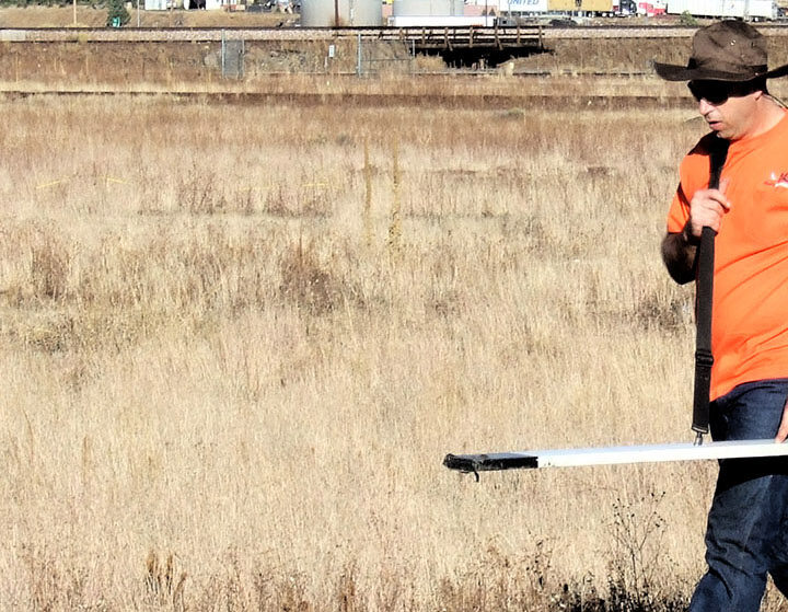 Person in orange shirt and hat walking in field, carrying a tripod and equipment.