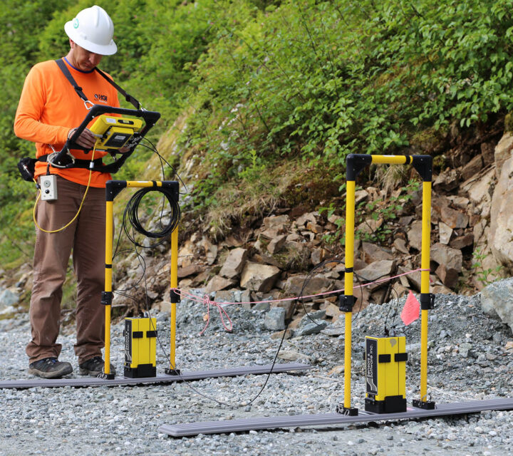 Worker using geophysical equipment on a gravel path near rocky hillside.