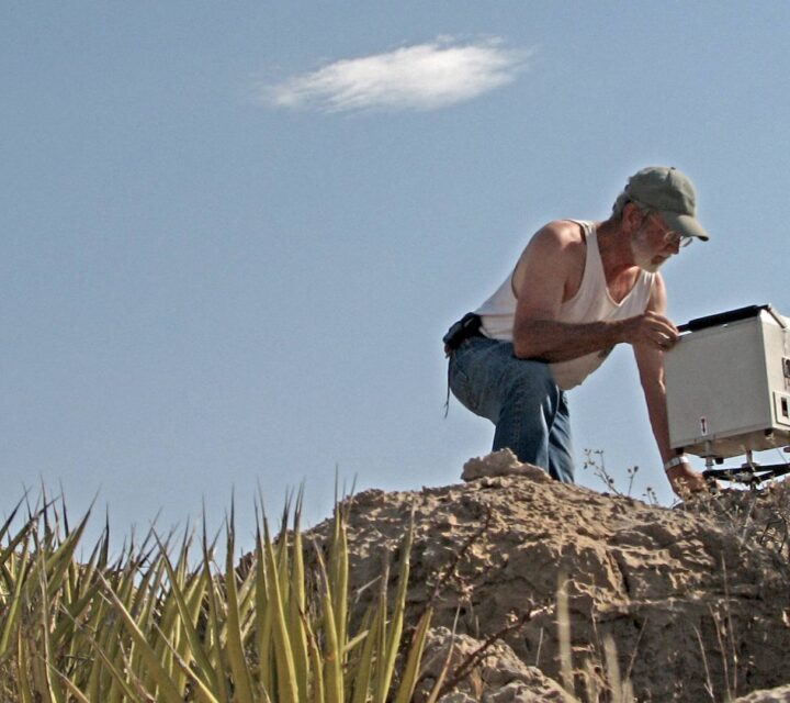 Man in cap setting up equipment on rocky terrain with clear sky background.