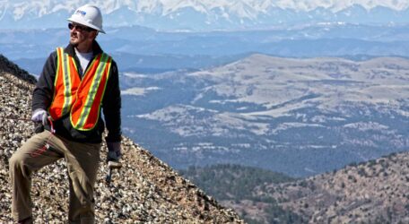 Worker in safety gear stands on rocky terrain with mountains in the background.