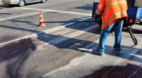 Worker applying asphalt sealant on a city street with a traffic cone nearby.