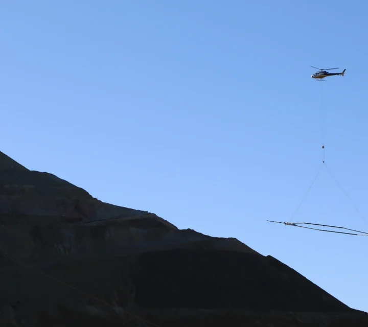 Helicopter conducting airborne electromagnetic method survey over mountainous terrain to map deep subsurface conductivity variations.