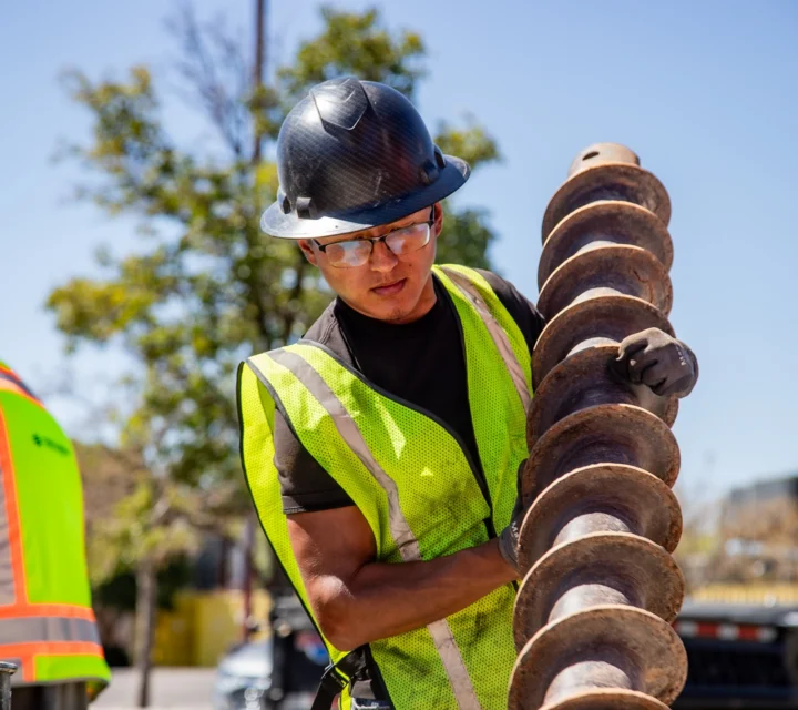 Construction worker in safety gear inspecting a large drill bit outdoors.