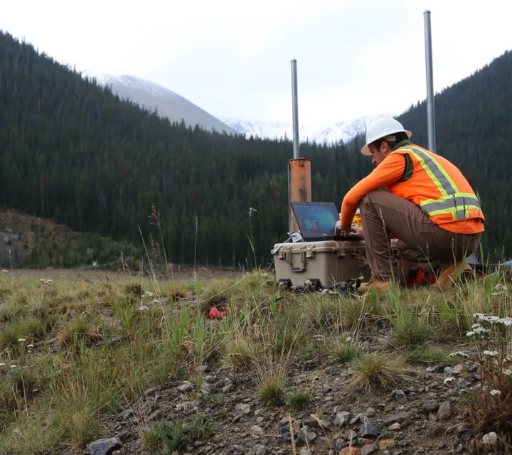 Geophysical technician conducting a bedrock depth mapping survey in a mountainous area, using field equipment and a laptop to evaluate subsurface variability and reduce structural risk.