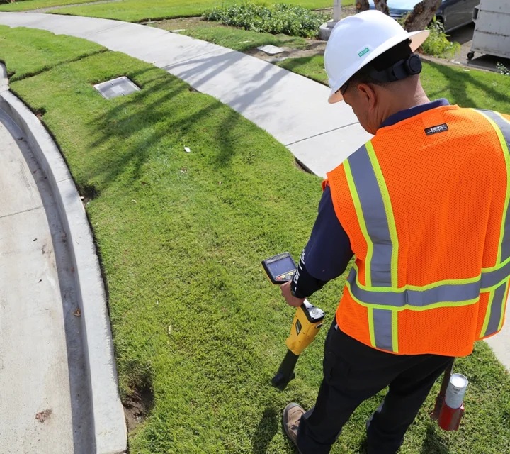 Worker in safety gear using a tool to scan underground on a grassy sidewalk.