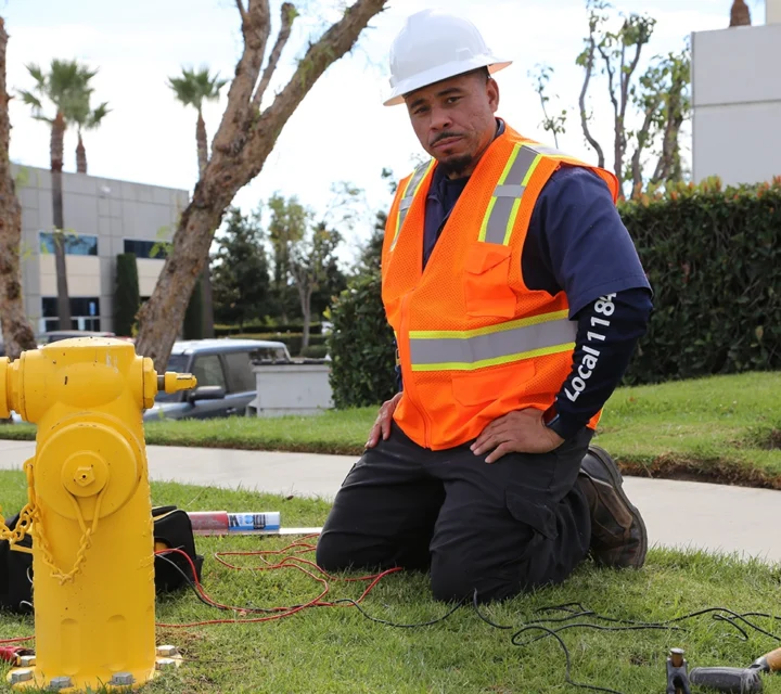Worker in safety gear kneeling near a yellow fire hydrant, with tools and wires on the ground.