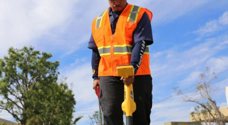 Worker in safety gear using a metal detector outdoors.
