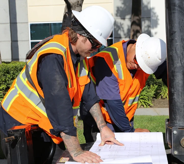 Two construction workers in safety gear reviewing blueprints at a worksite.