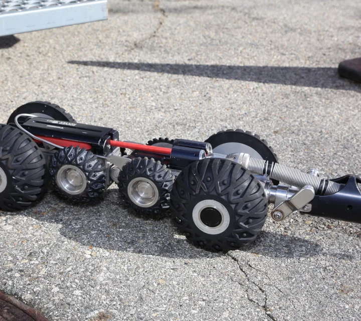 A wheeled robotic vehicle with large tires on concrete, near an orange traffic cone.