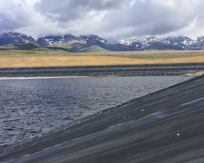 Reservoir with black lining, surrounded by hills and distant snowy mountains under a cloudy sky.