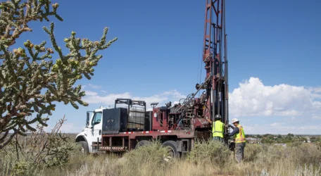 Workers operate a drilling rig in a desert landscape under a clear sky.