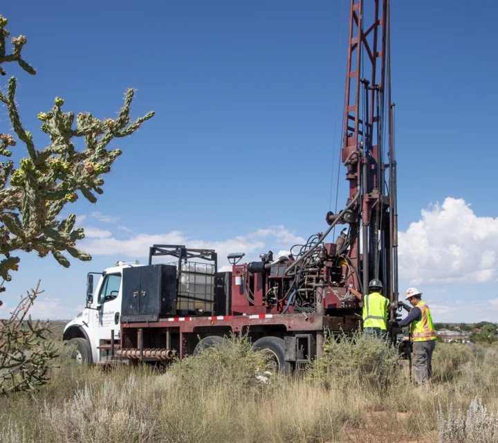 Workers operate a drilling rig in a desert landscape under a clear sky.