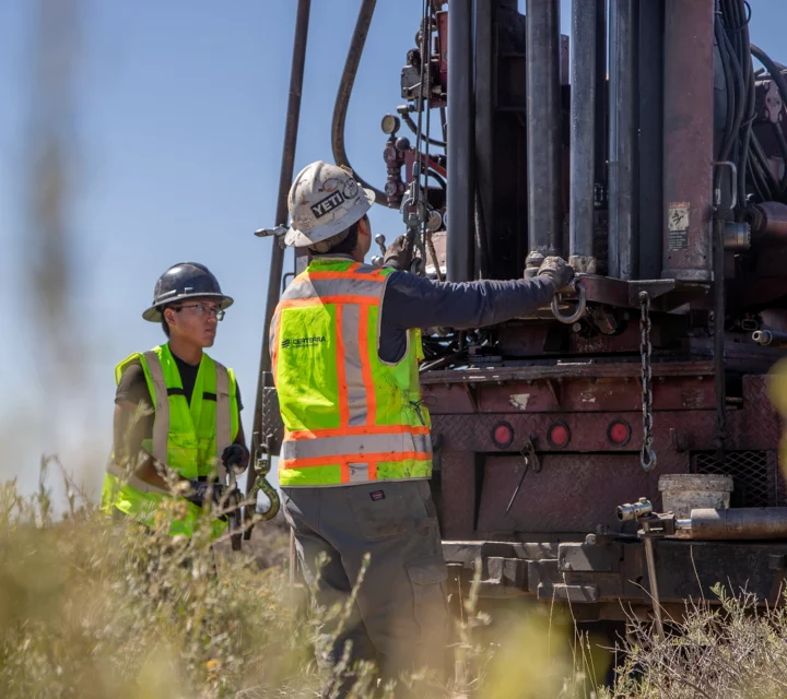 Two workers in safety gear operate heavy machinery outdoors in a field.