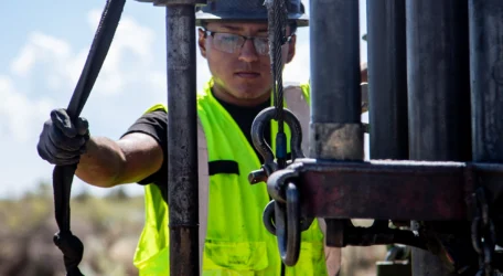 Worker in a hard hat and neon vest operates drilling equipment outdoors.