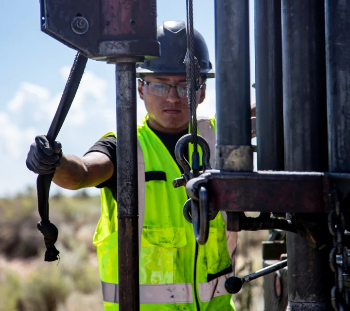 Worker in a hard hat and neon vest operates drilling equipment outdoors.