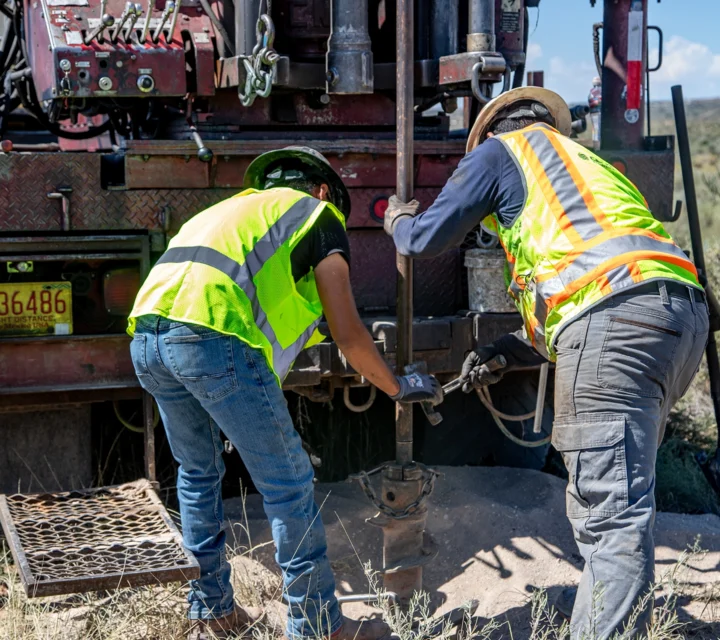 Two workers in safety vests operate a drilling rig in an open field.