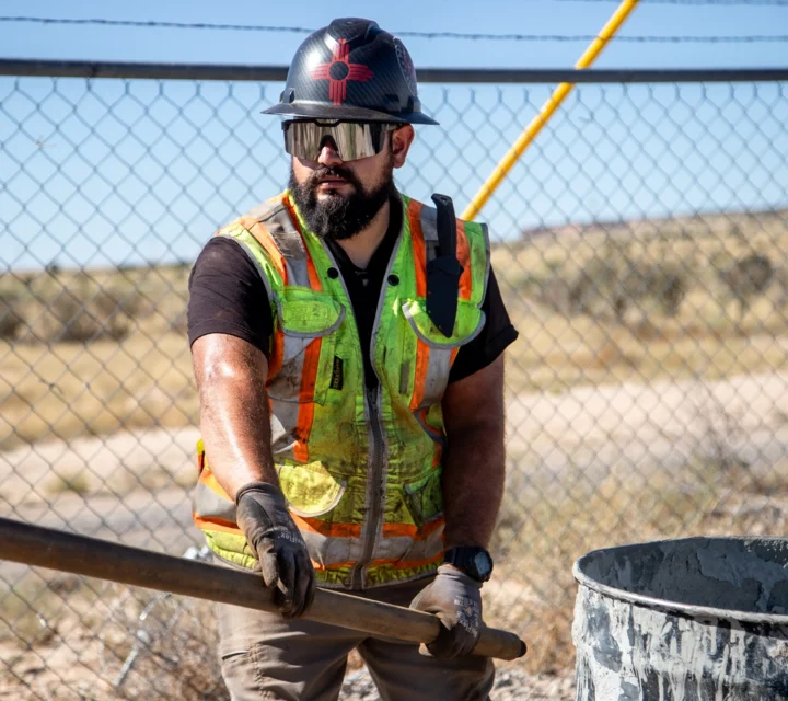 Construction worker with beard and helmet holding a metal pole near a fence on a sunny day.