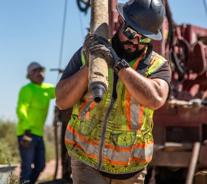 Construction worker carrying a large pipe, wearing safety gear and reflective vest, with a colleague in the background.