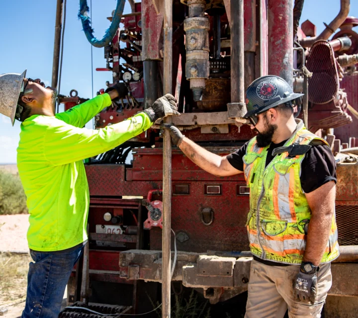 Two workers in safety gear operate a large industrial drilling machine outdoors.