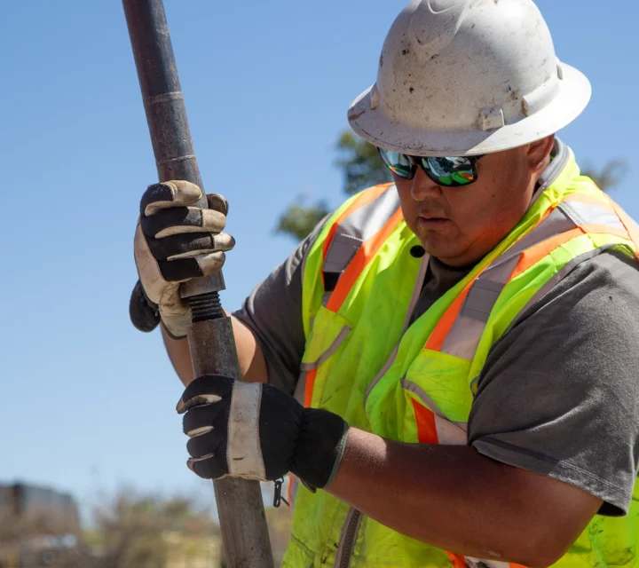 Construction worker in safety gear and helmet, holding a metal pole under a clear blue sky.