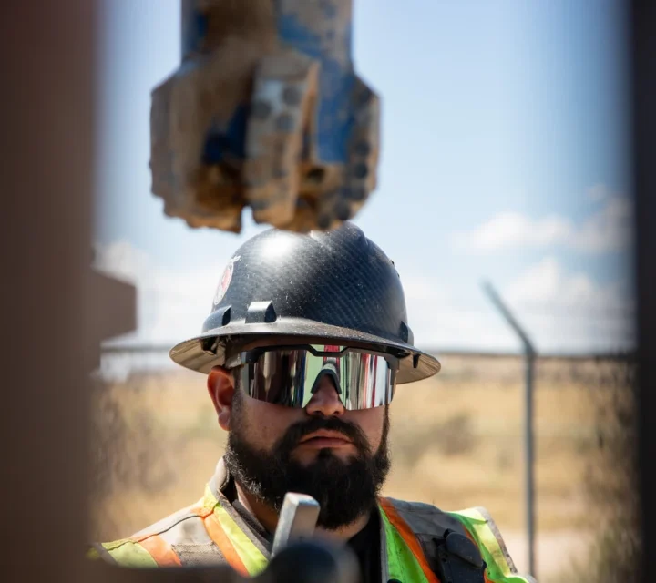 Construction worker in safety gear stands under heavy machinery, staring ahead, with a fence and sky in the background.