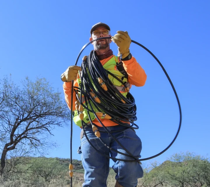 Worker with safety gear carrying coiled black cable in a rural area under clear blue sky.